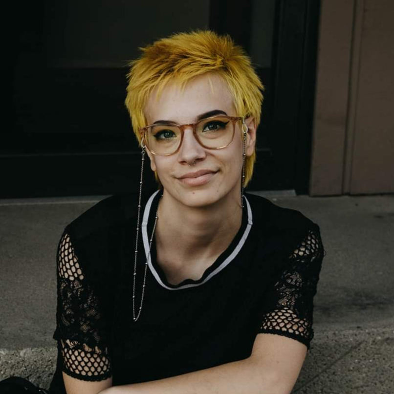Person with short yellow hair sitting on steps wearing a black top and checkered pants.